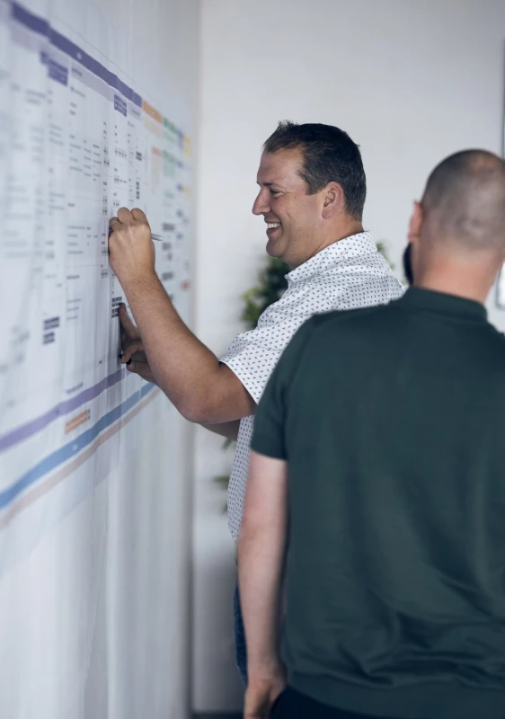 Two professionals evaluating a project timeline on a wall-mounted board in an office setting.